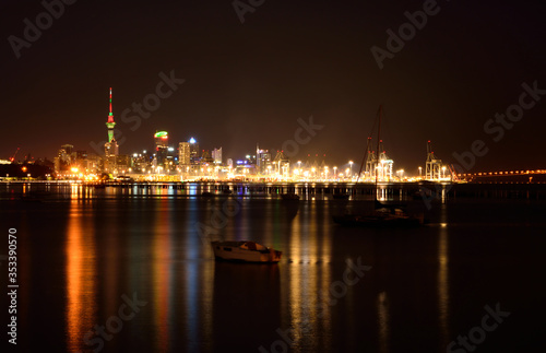 Midnight in Auckland. The lights of city downtown and the port reflected in Okahu bay with a small boat in the foreground. Soft focus