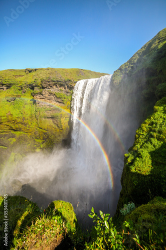 Icelandic waterfall Skogafoss with a rainbow and green moss. Icelandic waterfall. Skogafoss waterfall. Natural tourist attraction of Iceland. Summer landscape on a sunny day. Amazing landscape