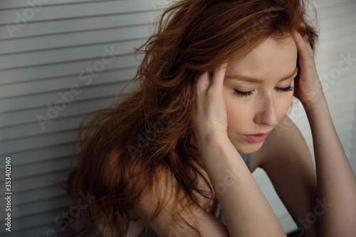 Redhead girl with freckles sits against a white wooden screen with closed eyes, fingers in her hair