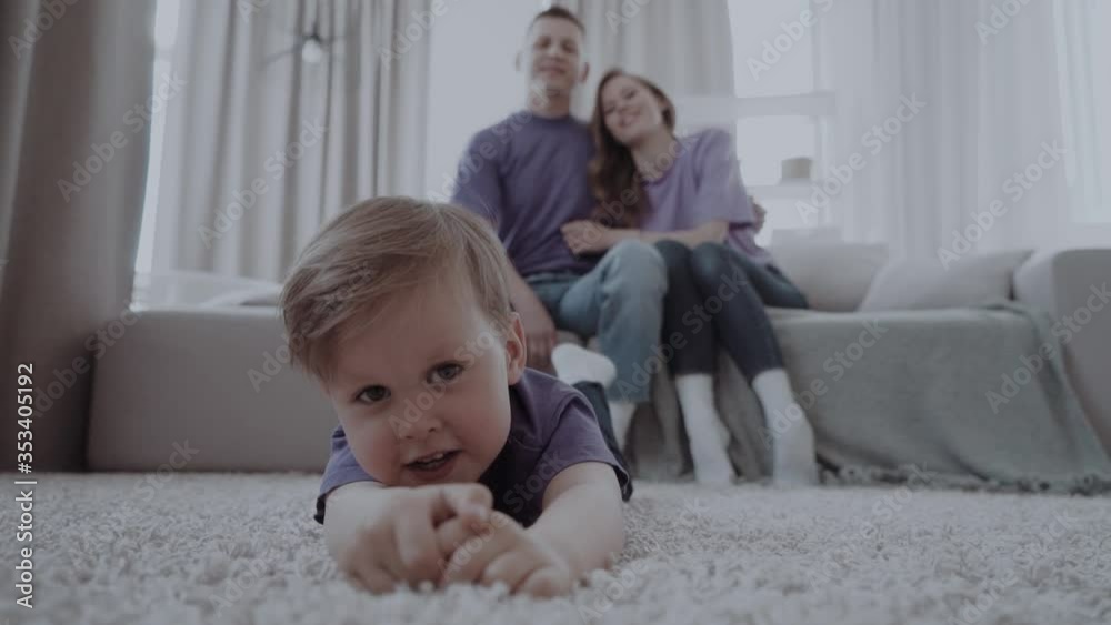 a small child is lying on the carpet in the living room while happy parents are sitting on the sofa on the background a small child is lying on the carpet in the living room while happy parents are sitting on the sofa on the background