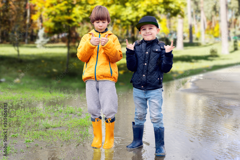 Two cheerfull friendly boys in rubber boots standing in puddle in park showing thumbs up gesture. Concept of social adaptation of autistic children. Friendship with peers. Full length portrait. Two cheerfull friendly boys in rubber boots standing in puddle in park showing thumbs up gesture. Concept of social adaptation of autistic children. Friendship with peers. Full length portrait.