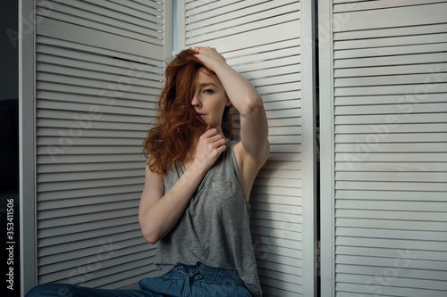 Redhead girl with freckles in a T-shirt sits leaning on a white wooden screen, and looks into the frame, covering face with hair.