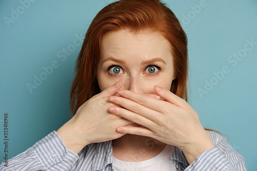 Red-haired girl is standing on a blue background in a shirt, looking at the camera with eyes wide open, covering mouth with hands