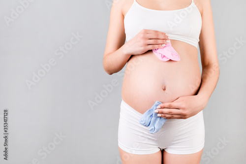 Close up of pregnant woman in white underwear holding baby pink and blue socks against her belly at gray background. Is it a boy or a girl? Waiting for twins. Child expecting concept. Copy space