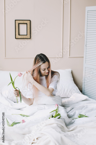 Romantic girl. Portrait of a girl in bed with natural flowers. Dreamy young woman with pink tulips resting in bedroom in morning