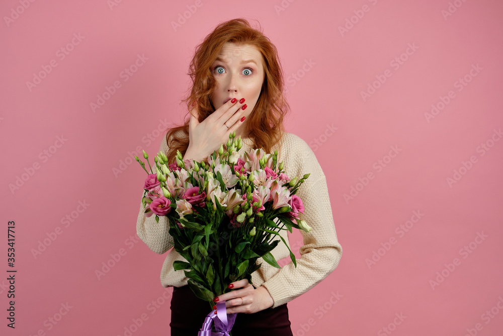 Cute red-haired girl on a pink background with a bouquet looks in surprise at the frame, closing her mouth with her hand