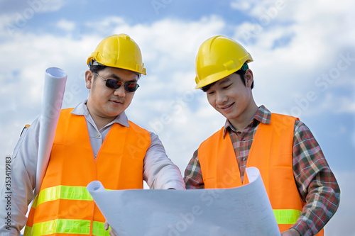 Two Asian engineers or technicians wear a yellow helmet with an orange reflective jacket and stand to hold a blueprint with a smile. Both consult the rooftop or construction site.