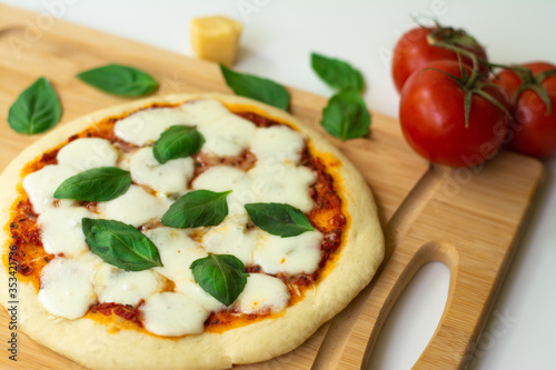 Homemade classic Italian Napoli pizza with tomato sauce, mozzarella cheese and basil leaves: pizza Napoletana. It is on a wooden cutting board surrounded by tomatoes, cheese and basil leaves