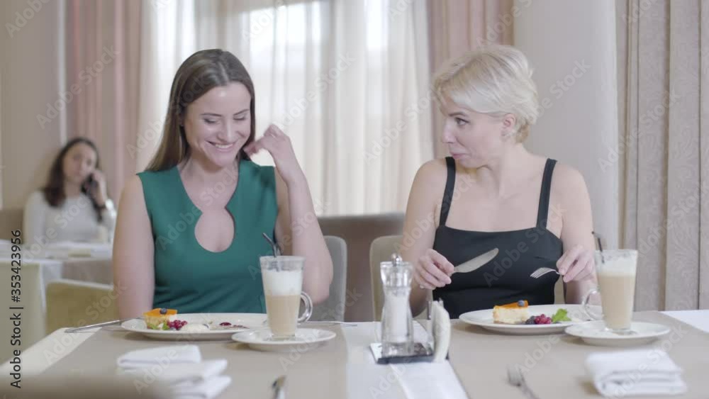 Positive female friends talking and eating dessert in restaurant. Portrait of two young women resting in cafe. Blond and brunette elegant ladies tasting sweet food. Leisure, lifestyle, friendship, joy Positive female friends talking and eating dessert in restaurant. Portrait of two young women resting in cafe. Blond and brunette elegant ladies tasting sweet food. Leisure, lifestyle, friendship, joy