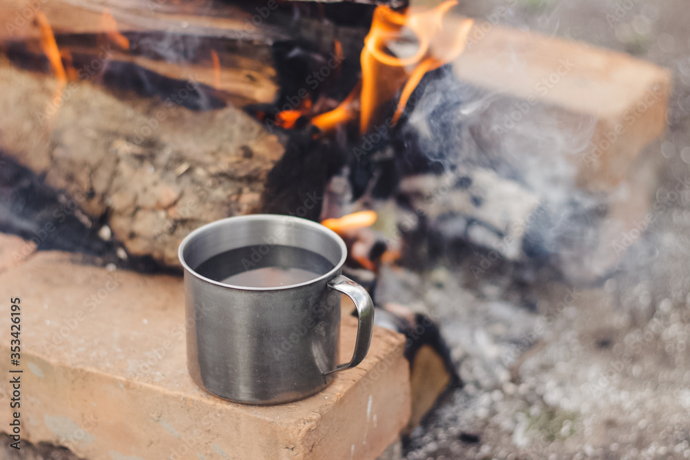Metal cup of coffee water standing on the orange clay brick near the flame bonfire with firewood and smoke in spring. Outdoor activity bbq. Metal cup of coffee water standing on the orange clay brick near the flame bonfire with firewood and smoke in spring. Outdoor activity bbq.