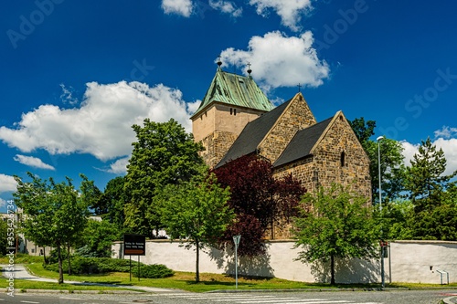 Kyje, Prague / Czech Republic - May 27 2020: View of the Church of Saint Bartholomew made of stone standing behind a wall. Green trees around. Sunny spring day with blue sky and white clouds.