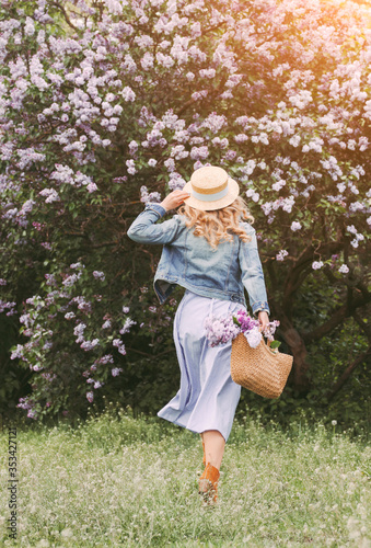 Back view cheerful carefree attractive blonde girl in denim jacket and dress relax, have fun in countryside sunny park. Happy beautiful young hipster woman run on blossom lilac garden. Freedom, peace