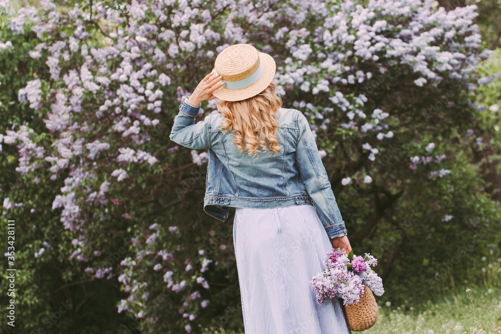 Back view stylish young beautiful blonde woman in denim jacket, dress, straw hat posing on blossom lilac garden. Happy attractive hipster girl walk, have fun in countryside park. Enjoy summer nature