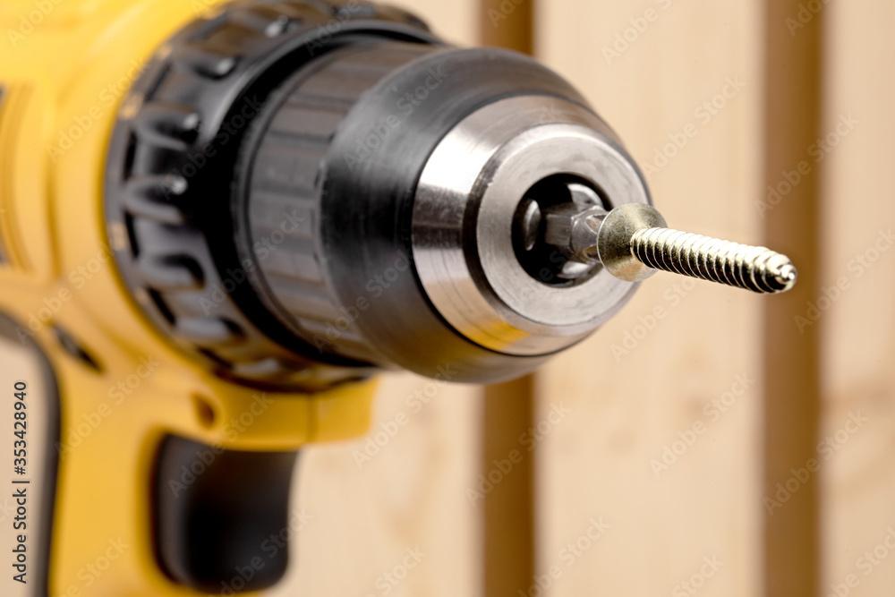 closeup of a yellow battery operated drill with screwdriver bit and wood screw pointing at the camera in a construction environment