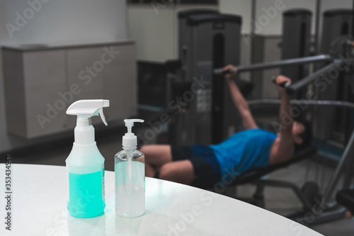 Close up spray bottle of blue disinfectant and alcohol gel with blurred background of Asian man workout in gym. Concept for prevention and protection of Corona virus Covid 19 in fitness center.