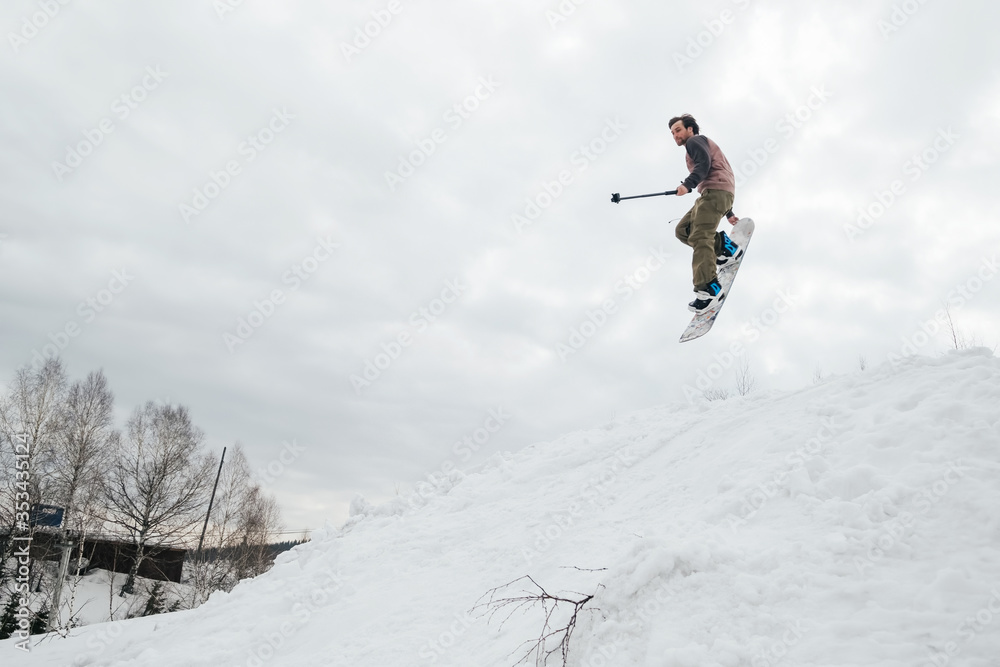 Snowboarder jumping from kicker in winter day holding action camera and shooting himself