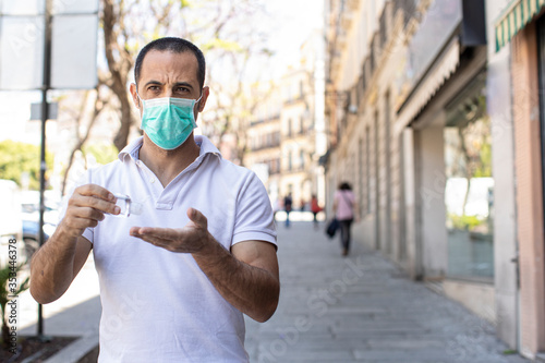 Uomo moro con polo bianco e mascherina chirurgica si disinfetta le mani cin un gel antibatterico  fermo in un marciapiede  photo