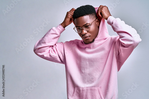 Close up of young african american man in pink streetwear hoodie with hood on head posing isolated on grey background in studio.