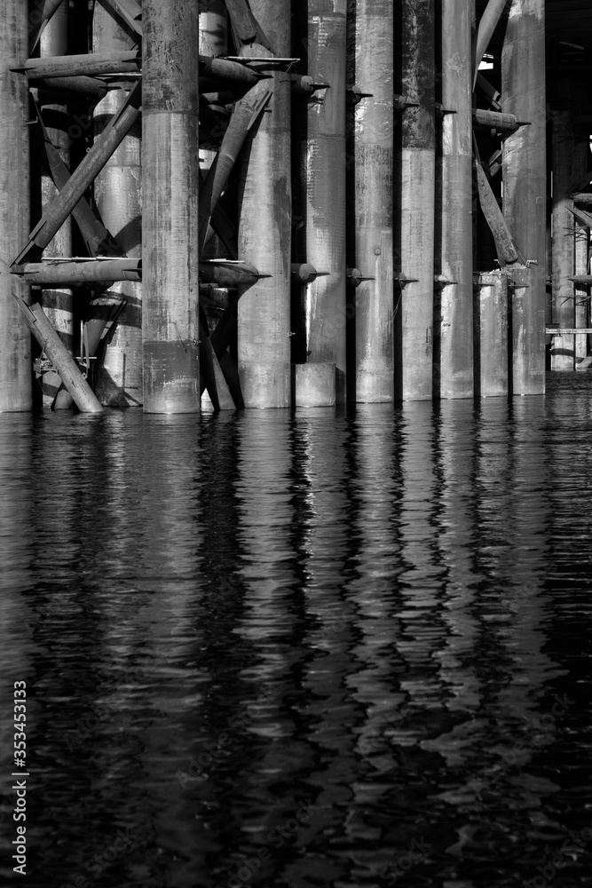 The rusty base of the bridge lowers and is reflected in the water. A black and white vertical industrial photo was made for your urban design.