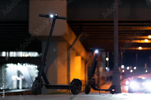 Black Electric Kick Scooters With Turn On Headlights at Cityscape at Night Time. Kick Scooters Near Passing By Cars on Urban Background photo
