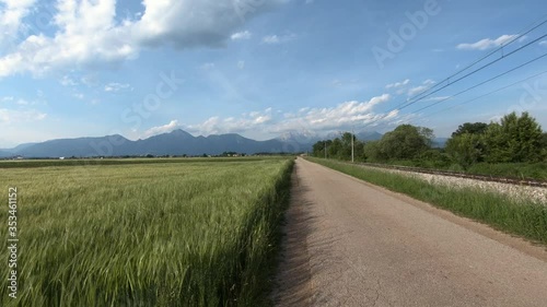 Walking on countryside narrow asphalt road. Farming field with growing vegetation. Railroad tracks on right side. Flatland basin. Alps mountains in background. Wide shot, forward dolly moving photo