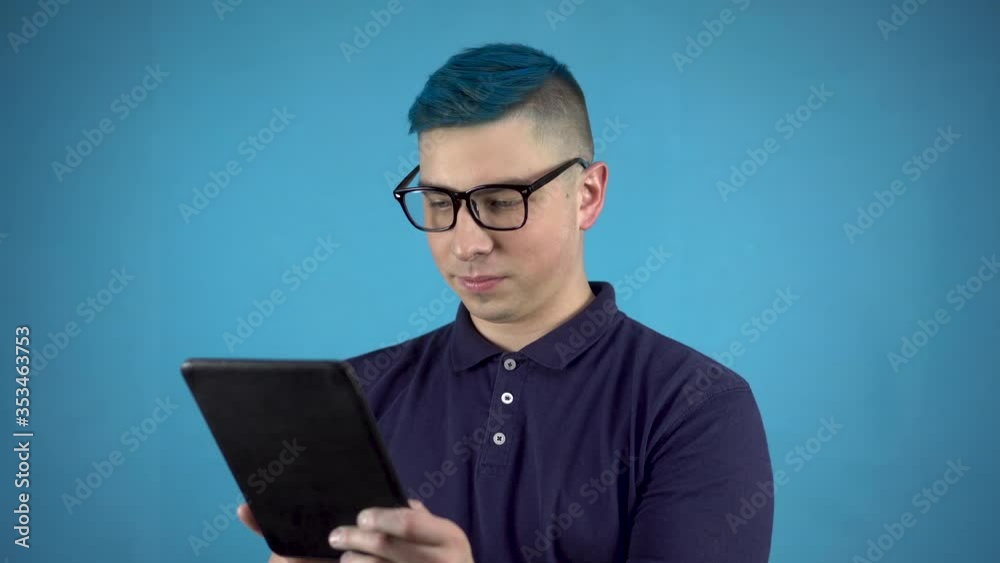Young man in glasses with blue hair with a tablet in his hands. Alternative man is writing in a tablet on a blue background. Young man in glasses with blue hair with a tablet in his hands. Alternative man is writing in a tablet on a blue background.
