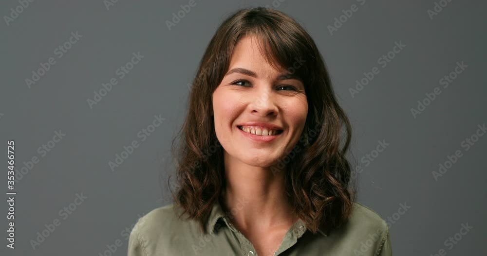 Close up of joyful pretty Caucasian young woman with smile on face indoor. Beautiful cheerful dark-haired female in good mood standing on grey background in studio and smiling. Portrait concept Close up of joyful pretty Caucasian young woman with smile on face indoor. Beautiful cheerful dark-haired female in good mood standing on grey background in studio and smiling. Portrait concept