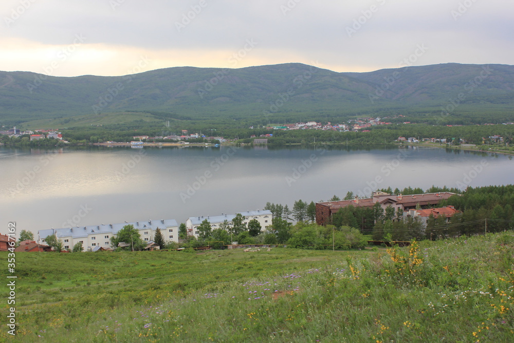 Obraz premium Great panoramic view. A lagoon with a clean mountain lake in the midst of majestic mountains in a haze of fog. Green grass. Apartments around the lake