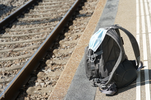  backpack with a n95 face mask attached on it, on a train station platform with rails in the background, transport at coronavirus covid-19 time with protection measures to prevent spread of infection
