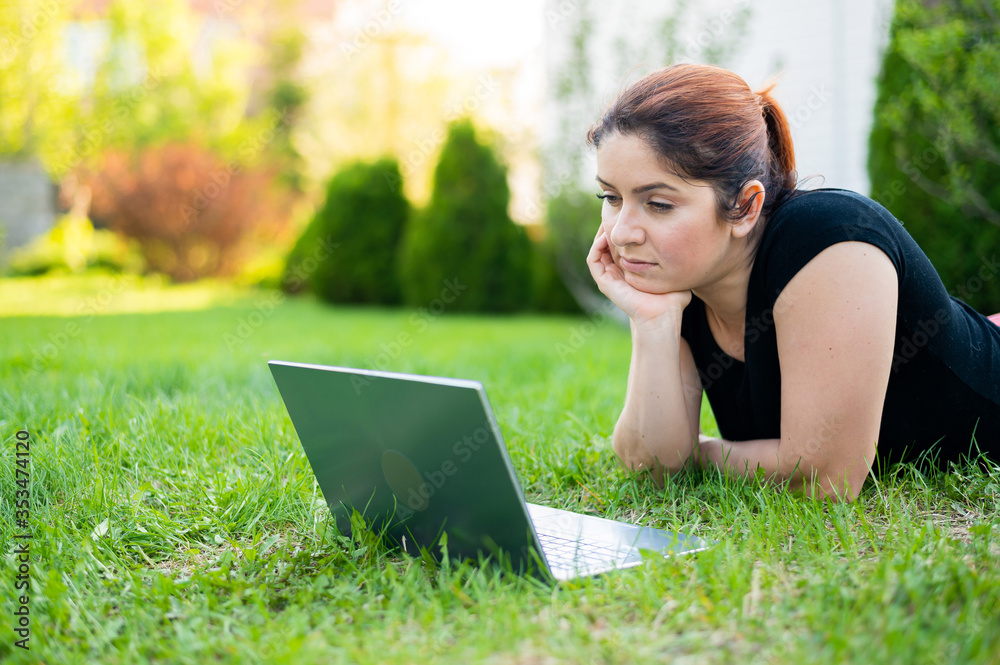 A woman lies on her stomach on the lawn at the cottage and uses on a laptop. A focused female freelancer works remotely outdoors. The girl is studying on the computer. A woman lies on her stomach on the lawn at the cottage and uses on a laptop. A focused female freelancer works remotely outdoors. The girl is studying on the computer.