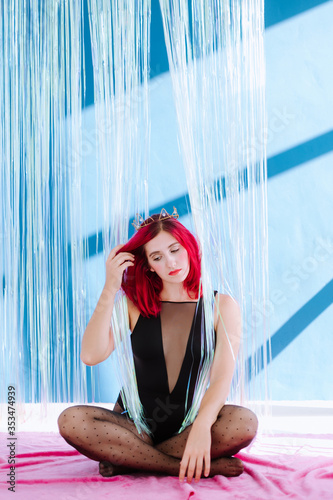 Full body vertical frame with young woman with red appearance in a black body sits on in the middle of shiny curtain against a blue wall and with a bored expression corrects hair withhand looking down photo