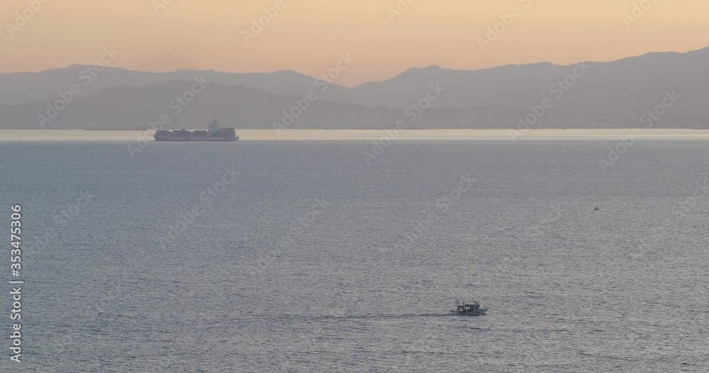 a small private boat and a big freighter transport ship on the quiet sea on the coast of Sardinia Italy at sunset with mountains in the background a small private boat and a big freighter transport ship on the quiet sea on the coast of Sardinia Italy at sunset with mountains in the background