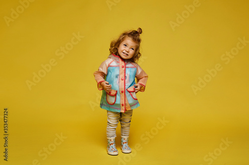 Cute little girl in a modish raincoat and rubber boots isolated at the yellow background. Child fashion. Copy space