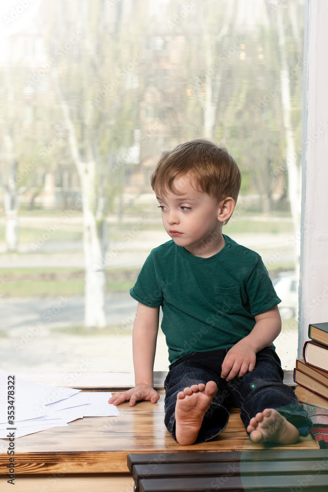 a three-year-old boy sits with a sad look on the windowsill. home leisure during quarantine and self-isolation. books and pencils nearby. view of the street and trees from the window. a three-year-old boy sits with a sad look on the windowsill. home leisure during quarantine and self-isolation. books and pencils nearby. view of the street and trees from the window.