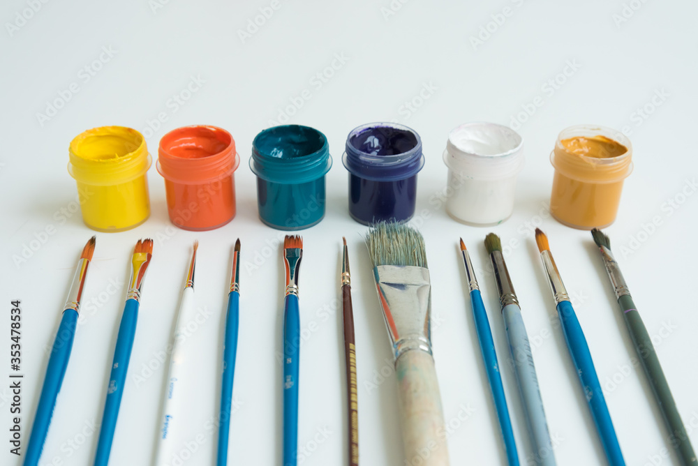 Jars of gouache paint in different colors. Next to it are brushes of various sizes. Artist's kit. Side view. White background. Hobbies and inspiration. Jars of gouache paint in different colors. Next to it are brushes of various sizes. Artist's kit. Side view. White background. Hobbies and inspiration.