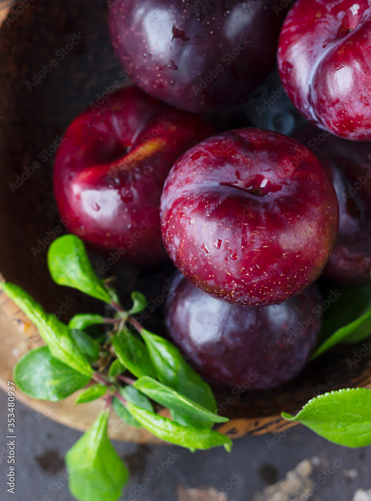 Obraz premium Ripe juicy purple plums with drops in wooden bowl with leaves, dark brown concrete background. Close up.