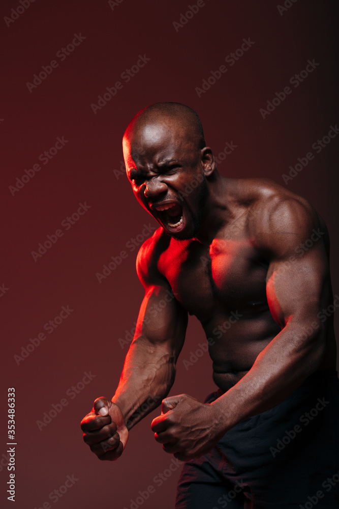 Sports shot with a red light of a dark-skinned muscular bodybuilder showing naked torso muscles and screaming