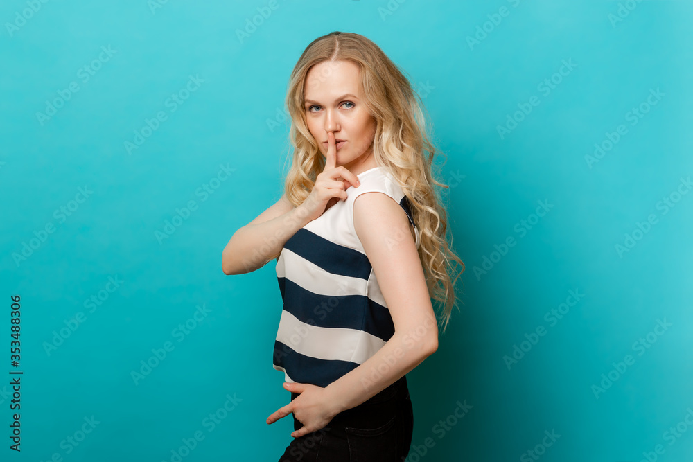 Portrait of a beautiful woman with long, lush hair in a striped t-shirt, holding her index finger on her lips, standing isolated against a blue background Portrait of a beautiful woman with long, lush hair in a striped t-shirt, holding her index finger on her lips, standing isolated against a blue background