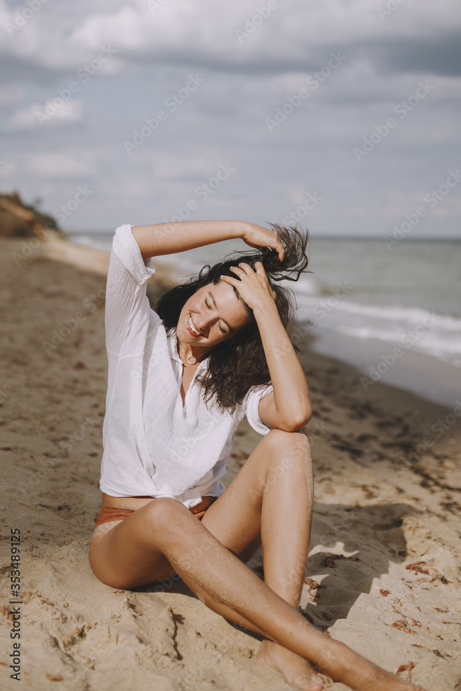 Happy boho girl in white shirt sitting on sunny beach. Carefree stylish woman smiling and relaxing on seashore. Summer vacation. Lifestyle authentic image. Mindfulness.