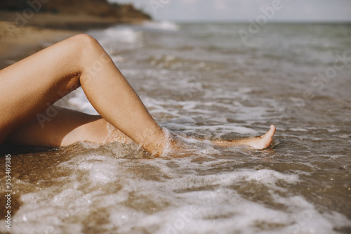 Legs in waves foam on beach closeup. Young woman relaxing on seashore, sitting on sandy beach with waves. Summer vacation. Carefree moment. Authentic image