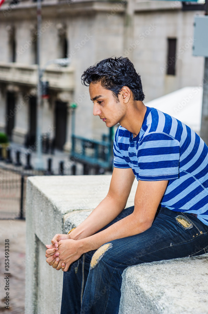 Lonely East Indian American teenager thinking outside in New York City, wearing blue line striped T shirt, broken fashionable jeans, sitting on street, looking down, sad, depressed, lost in thought..
