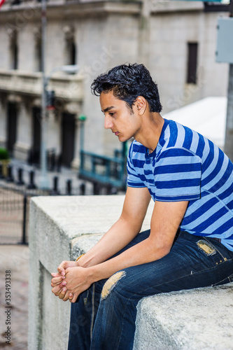 Lonely East Indian American teenager thinking outside in New York City, wearing blue line striped T shirt, broken fashionable jeans, sitting on street, looking down, sad, depressed, lost in thought..