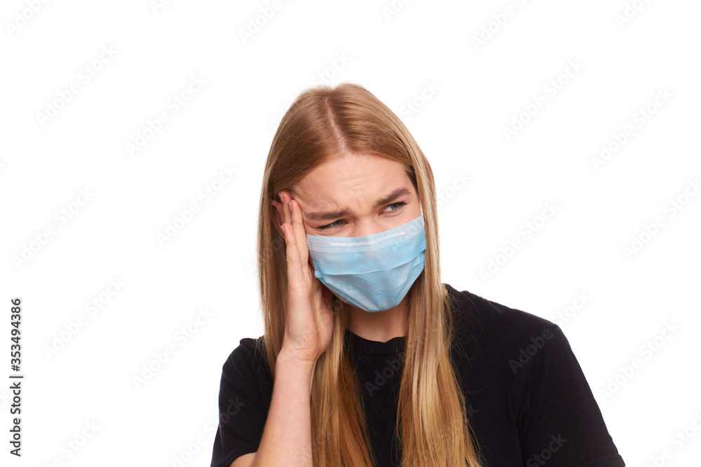 Woman wears protective white mask against virus, feels strong pain in temples, holding fingers to head. Young girl with long blonde dressed in black t-shirt Woman wears protective white mask against virus, feels strong pain in temples, holding fingers to head. Young girl with long blonde dressed in black t-shirt