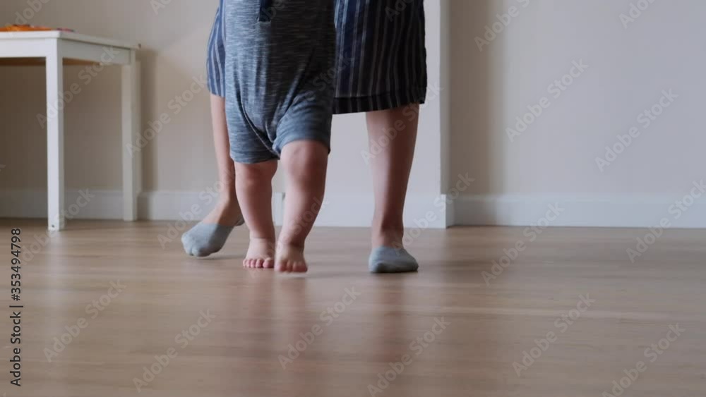 Little kid feet with mother walking on floor, close-up. Baby learning to walk at home. Baby first steps. Slow motion Little kid feet with mother walking on floor, close-up. Baby learning to walk at home. Baby first steps. Slow motion