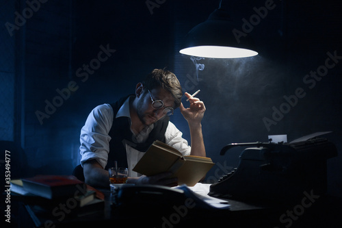 Medium sharp. Closeup portrait of a young writer who is sitting at a table with paper and glasses in his hand and a vintage typewriter. On the table is a glass of whiskey, an ashtray, books.