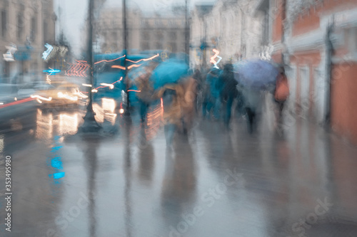 Rainy evening. Blurred silhouette of abstract silhouettes of people under umbrellas on the sidewalk near roadway, city life, traffic, illumination