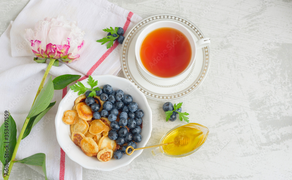 Tiny pancake cereal with blueberry, honey and tea for breakfast in the morning light and pink peony on table Tiny pancake cereal with blueberry, honey and tea for breakfast in the morning light and pink peony on table