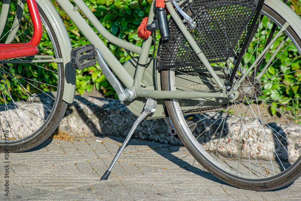 A gray bike with red trim stands on a backup on the sidewalk. Bicycle pedals close up.