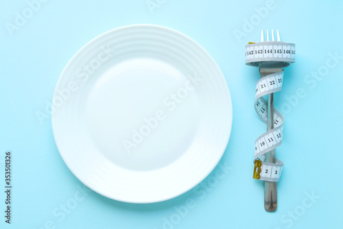 A white empty flat plate with a fork wrapped in measuring tape on a blue background. Diet and fasting. Weight loss. Selective focus . The view from the top