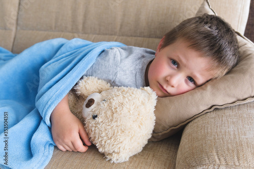 A sick or sad boy lies on a sofa under a blue blanket in an embrace with a teddy bear. Misunderstanding of parents, childhood depression. Copy space